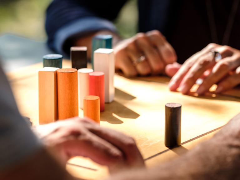 Zwei Menschen sitzen am Tisch, darauf ein Brett mit Holzelementen verschiedener Größe. Von den beiden Menschen sieht man nur die Hände.