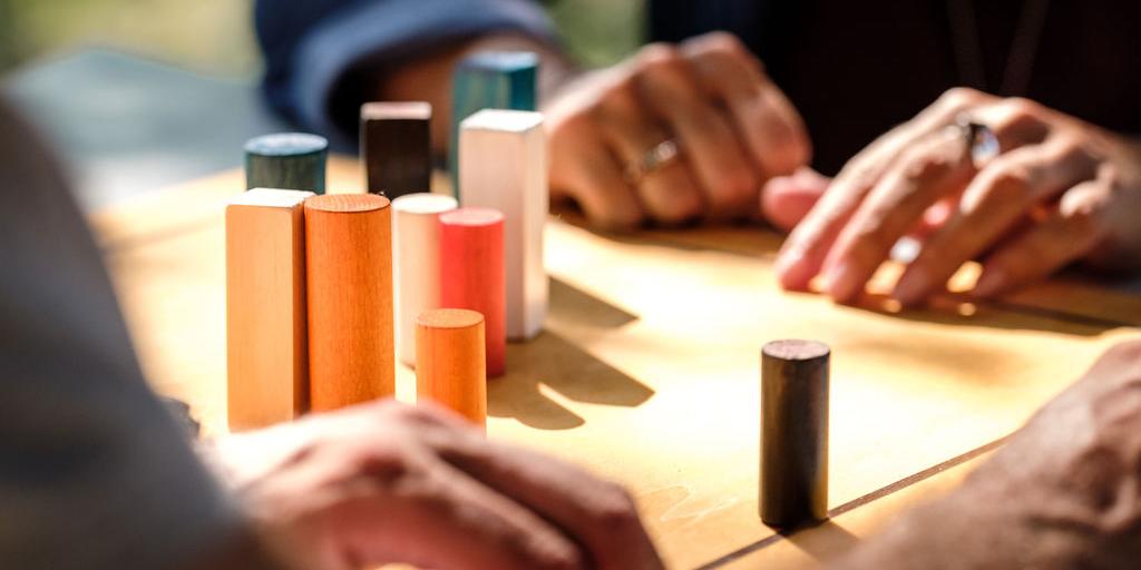 Zwei Menschen sitzen am Tisch, darauf ein Brett mit Holzelementen verschiedener Größe. Von den beiden Menschen sieht man nur die Hände.