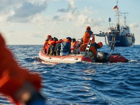 Szene aus dem Dokumentarfilm 'Route 4'. Geflüchtete auf einem zu kleinen Boot auf dem Meer, im Hintergrund ein Schiff.
