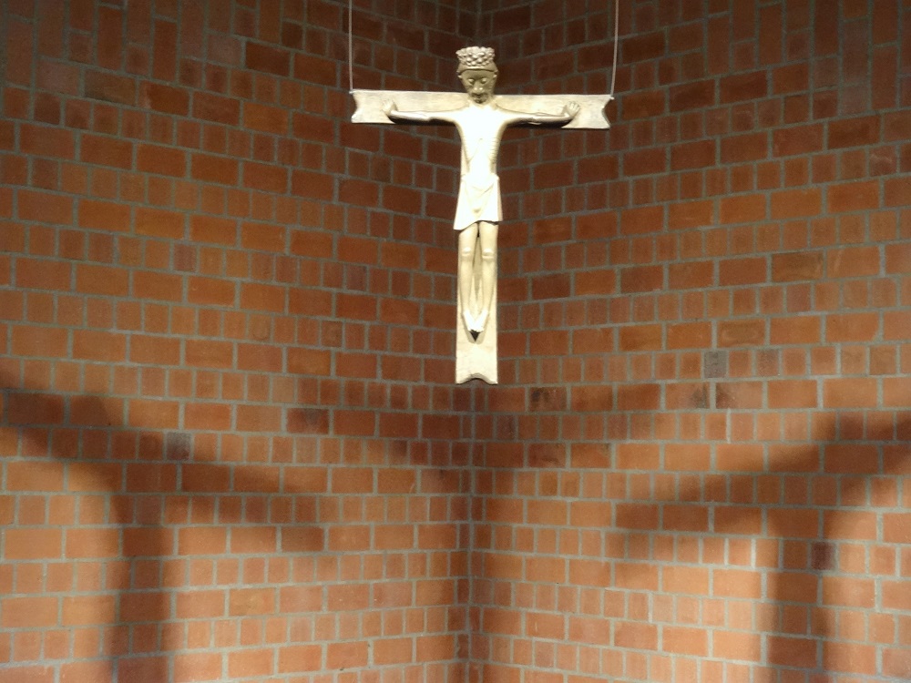 Ein Kreuz aus Metall hängt von einer Kirchendecke herab, man sieht einen dreifachen Schatten auf der Steinwand dahinter.