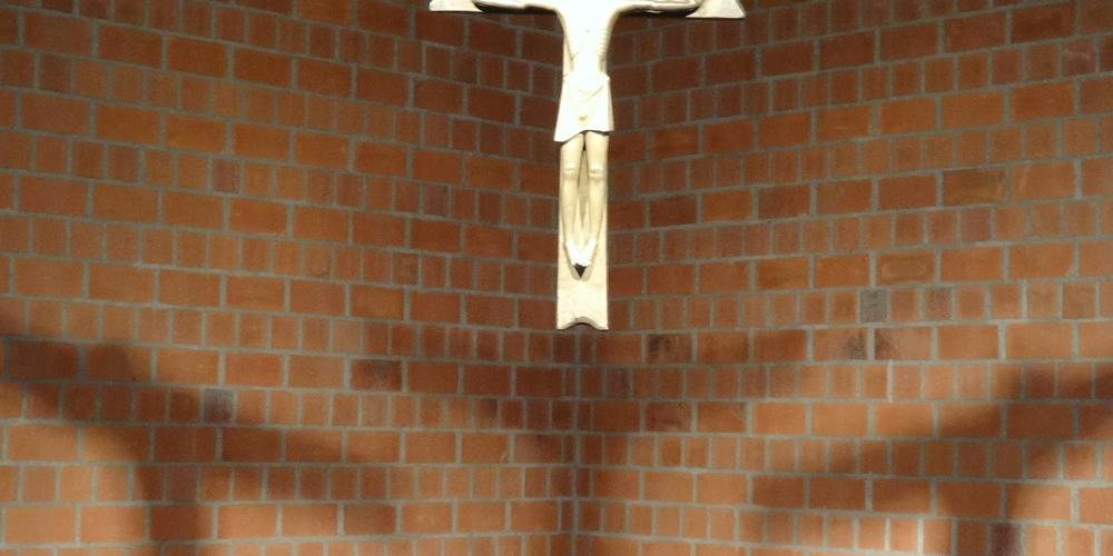 Ein Kreuz aus Metall hängt von einer Kirchendecke herab, man sieht einen dreifachen Schatten auf der Steinwand dahinter.