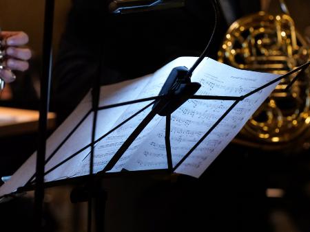 Notenblatt im Orchester (c) Peter Weidemann/Pfarrbriefservice.de Notenständer, auf dem Notenblätter liegen, von hinten fotografiert, dahinter im Ausschnitt zwei Musiker mit ihren Blasinstrumenten.