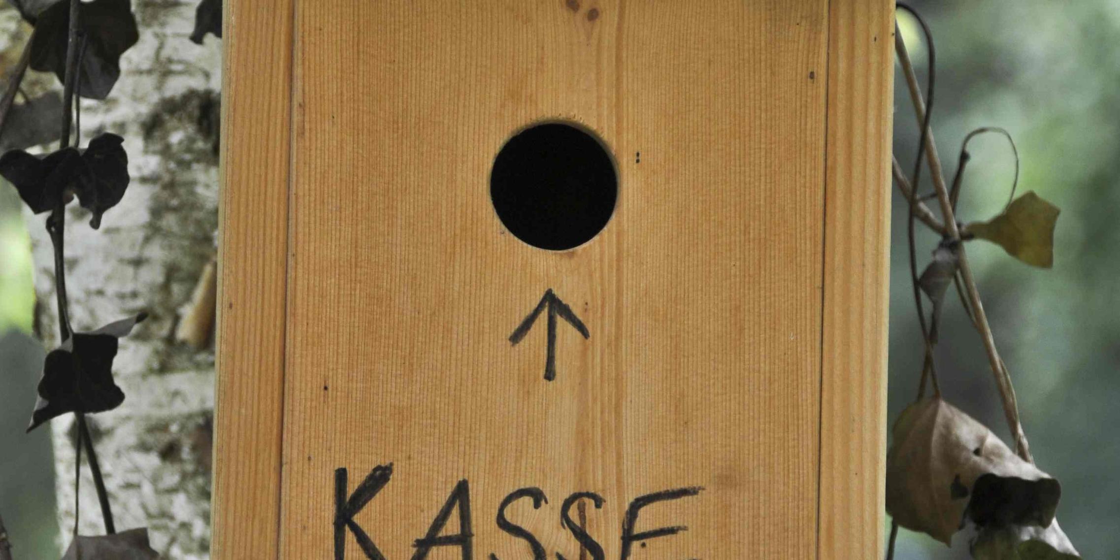 Ein Nistkasten aus Holz an einem Baum mit einem kleinen Schlupfloch, darunter die Aufschrift 'Kasse'