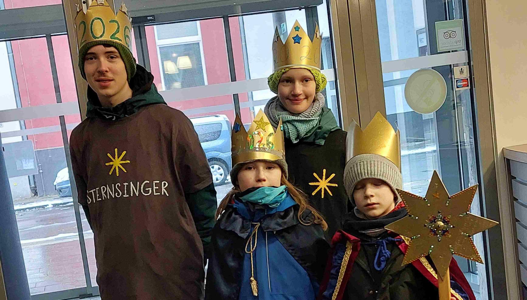 Vier Kinder, als Sternsinger mit Kronen und Stern, vor der Eingangstür der Akademie.