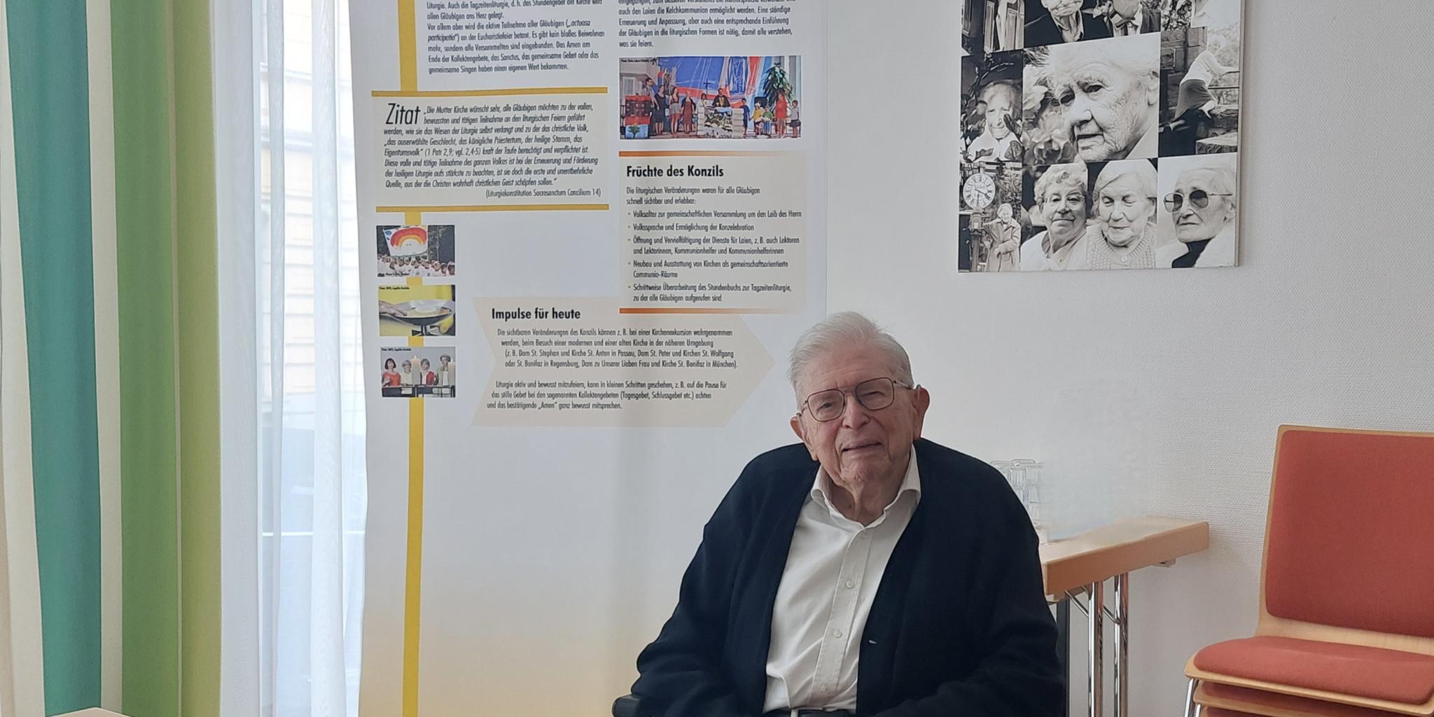 91-jähriger Priester im Rollstuhl vor einem Rollup mit Texten und Bildern zur Liturgiekonstitution des Zweiten Vatikanischen Konzils