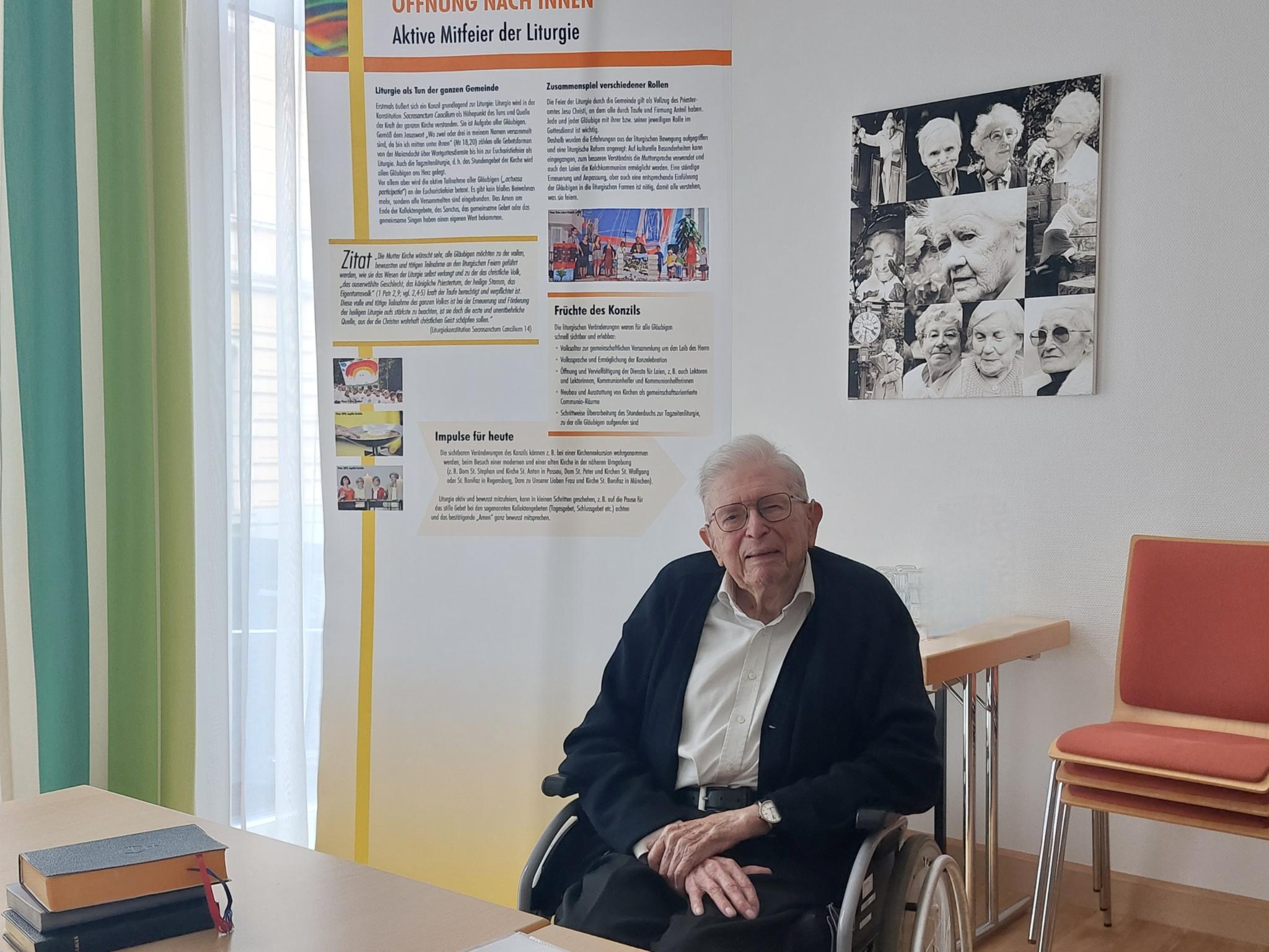 91-jähriger Priester im Rollstuhl vor einem Rollup mit Texten und Bildern zur Liturgiekonstitution des Zweiten Vatikanischen Konzils