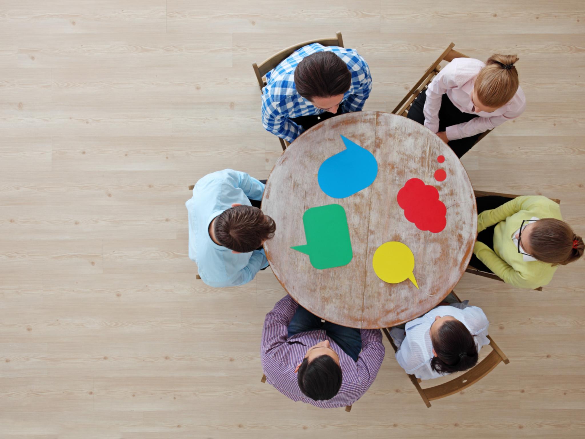 Menschen sitzen um einen Tisch, vor ihnen liegen Sprechblasen in unterschiedlicher Form.