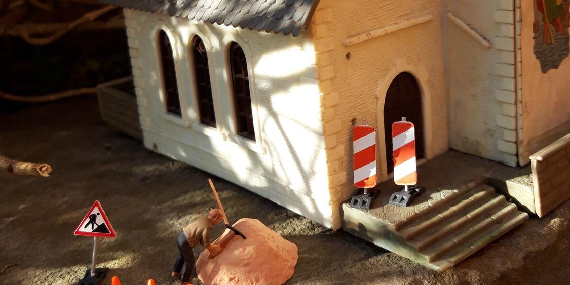 Vor einem Kirchengebäude im leichten Sonnenlicht ist eine Baustelle eingerichtet, ein Bauarbeiter arbeitet an einem Hügel mit Sand.