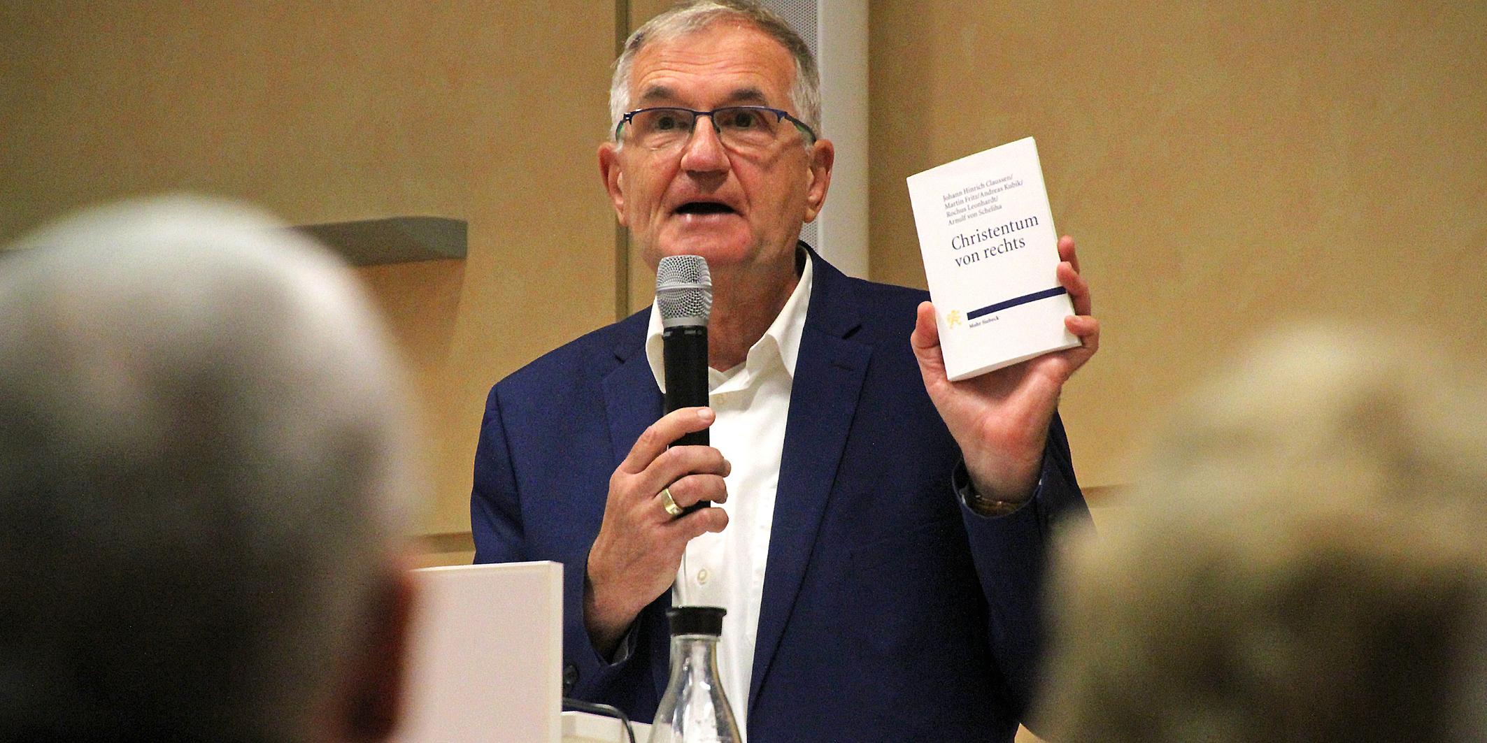 Andreas Püttmann, ein Journalist, hält ein Buch hoch und spricht ins Mikrophon