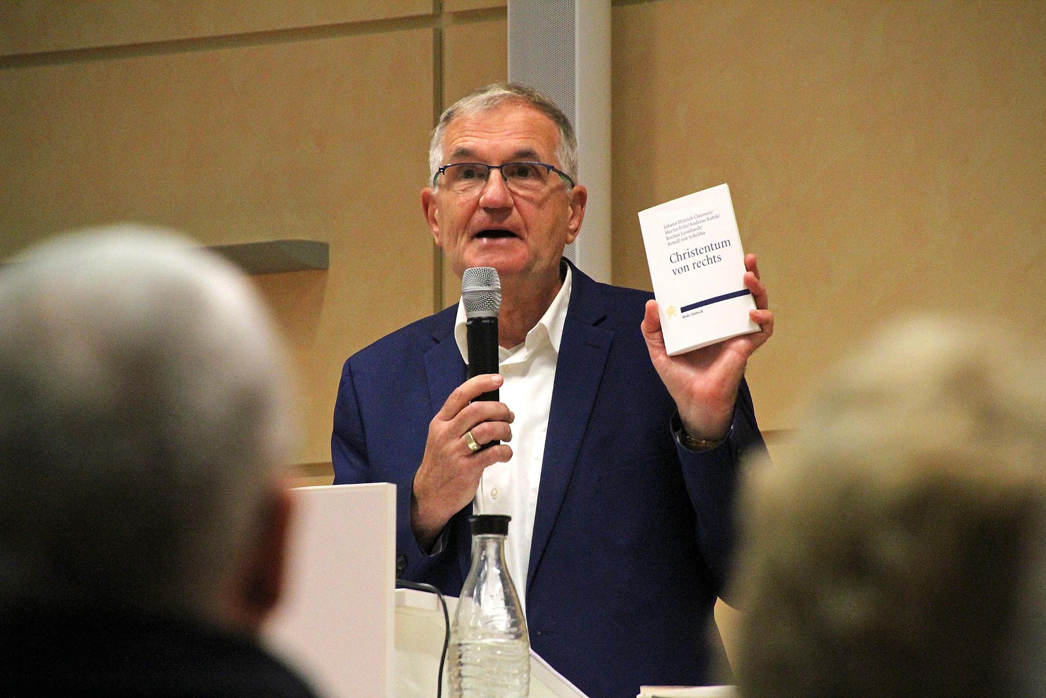 Andreas Püttmann, ein Journalist, hält ein Buch hoch und spricht ins Mikrophon