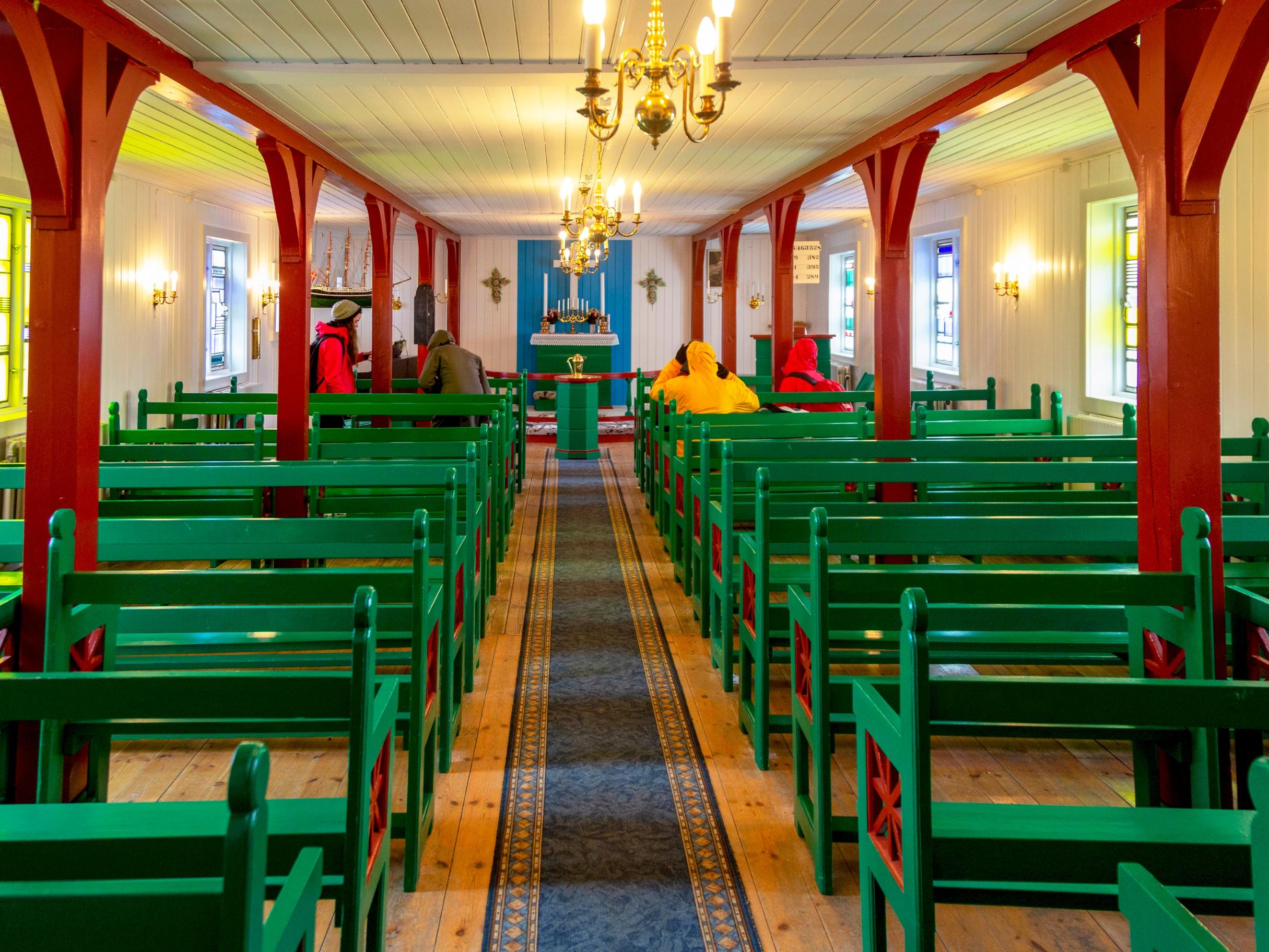 Das Innere einer lutherischen Kirche in Kulusuk auf Grönland, vornehmlich aus Holz gebaut, mit Bänken rechts und links des Mittelgangs. Seitlich sind Bleiglasfenster der Aachener Glasmalerin Maria Katzgrau zu sehen.