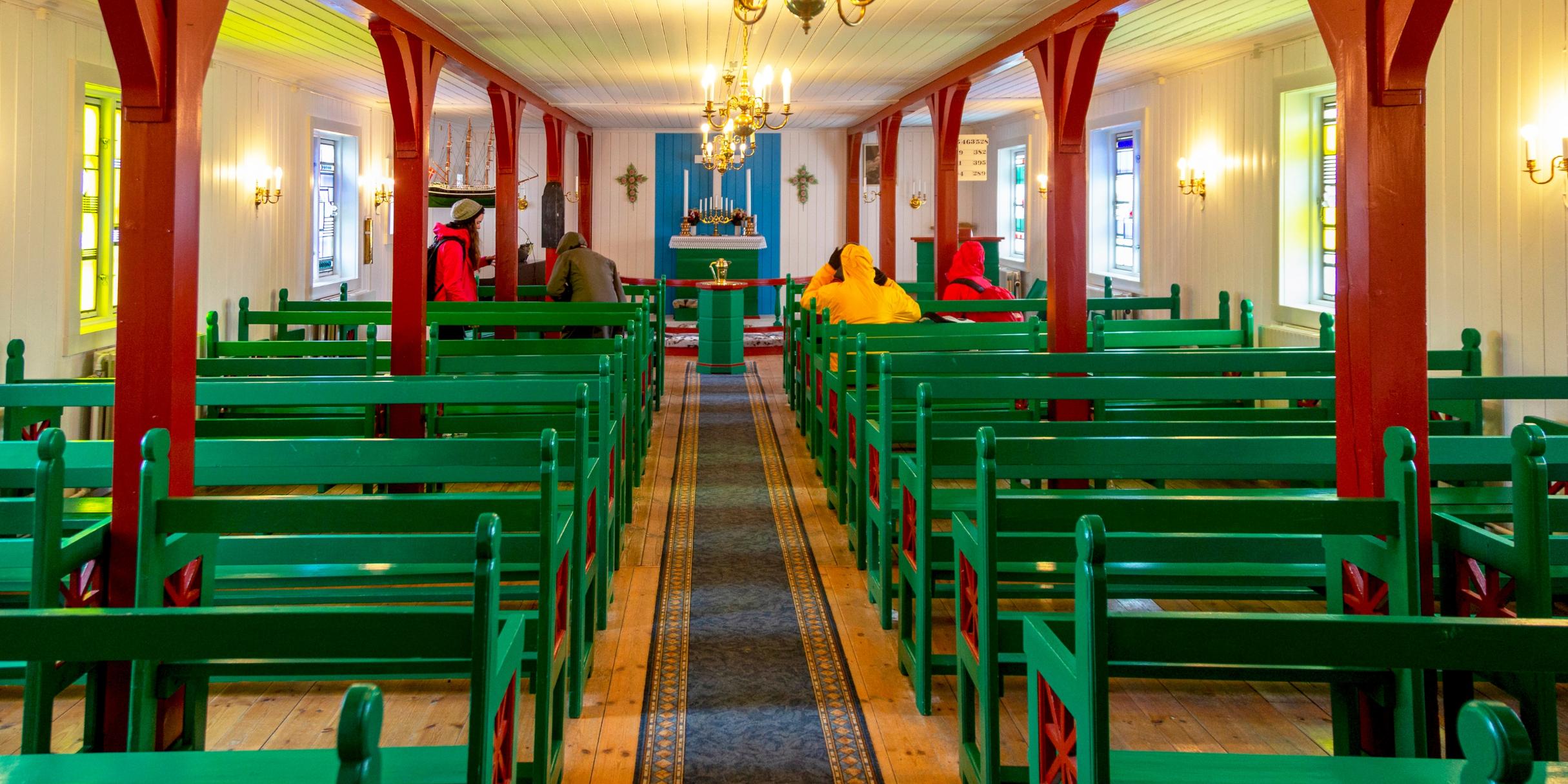 Das Innere einer lutherischen Kirche in Kulusuk auf Grönland, vornehmlich aus Holz gebaut, mit Bänken rechts und links des Mittelgangs. Seitlich sind Bleiglasfenster der Aachener Glasmalerin Maria Katzgrau zu sehen.