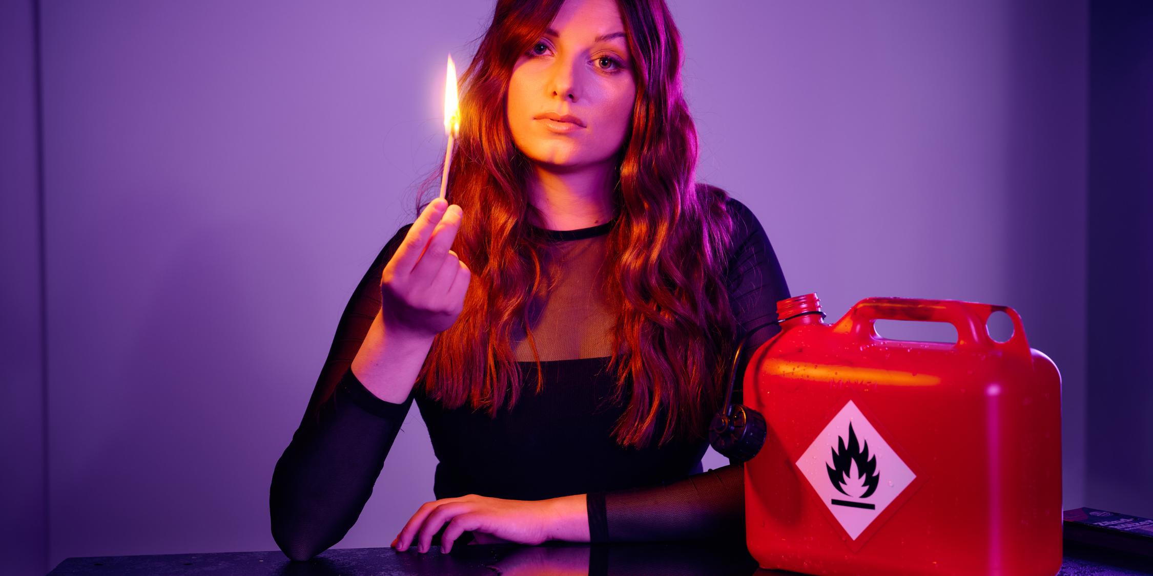 Die Kabarettistin Lara Ermer sitzt an einem Tisch. In einer Hand hält sie ein entzündetes Streichholz, neben ihr steht ein Behälter, der mit einem Warnaufkleber 'entflammbar' gekennzeichnet ist.