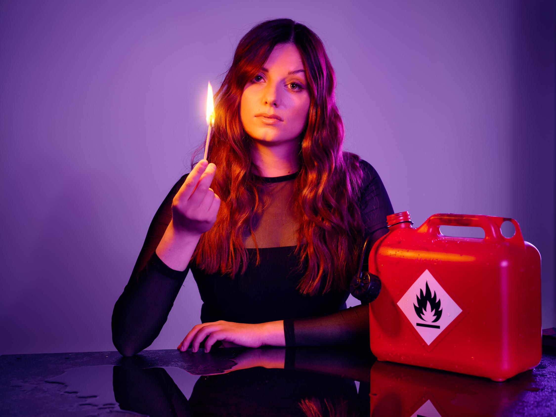 Die Kabarettistin Lara Ermer sitzt an einem Tisch. In einer Hand hält sie ein entzündetes Streichholz, neben ihr steht ein Behälter, der mit einem Warnaufkleber 'entflammbar' gekennzeichnet ist.