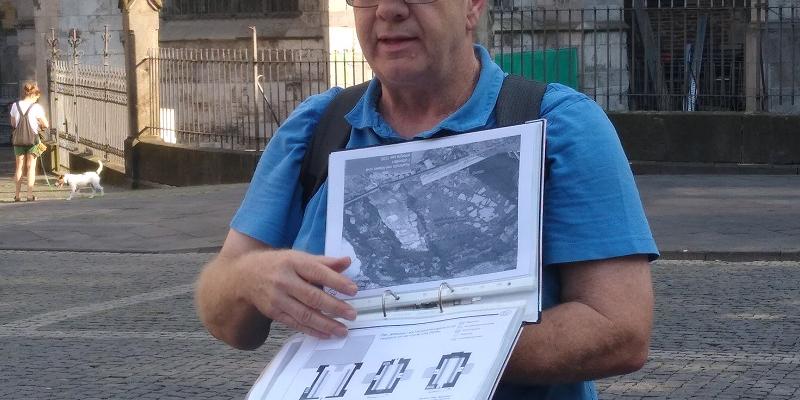 Ein Archäologe hält eine Mappe mit Einsteckhüllen in der Hand und zeigt ein Bild einer Ausgrabungssituation.