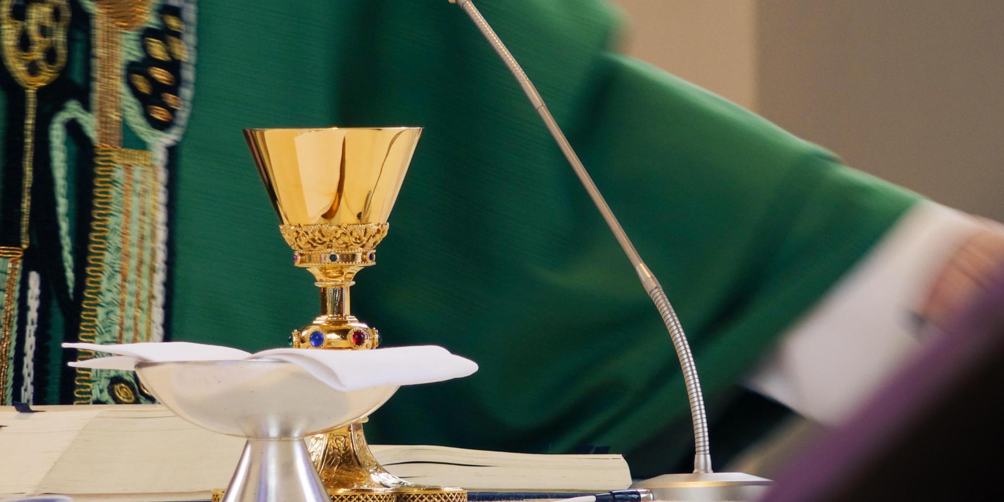 Auf dem Altar stehen Kelch und Patene, dahinter sieht man einen Teil eines Priesters im grünen Messgewand.