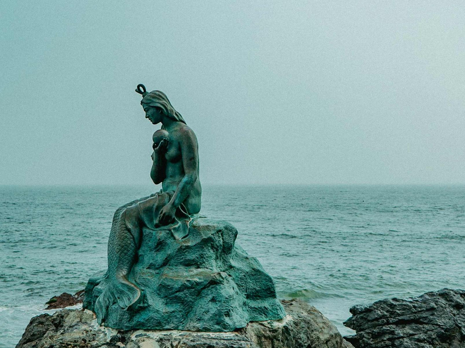 Skulptur einer Meerjungfrau, die traurig auf einem Stein sitzt und eine große Perle