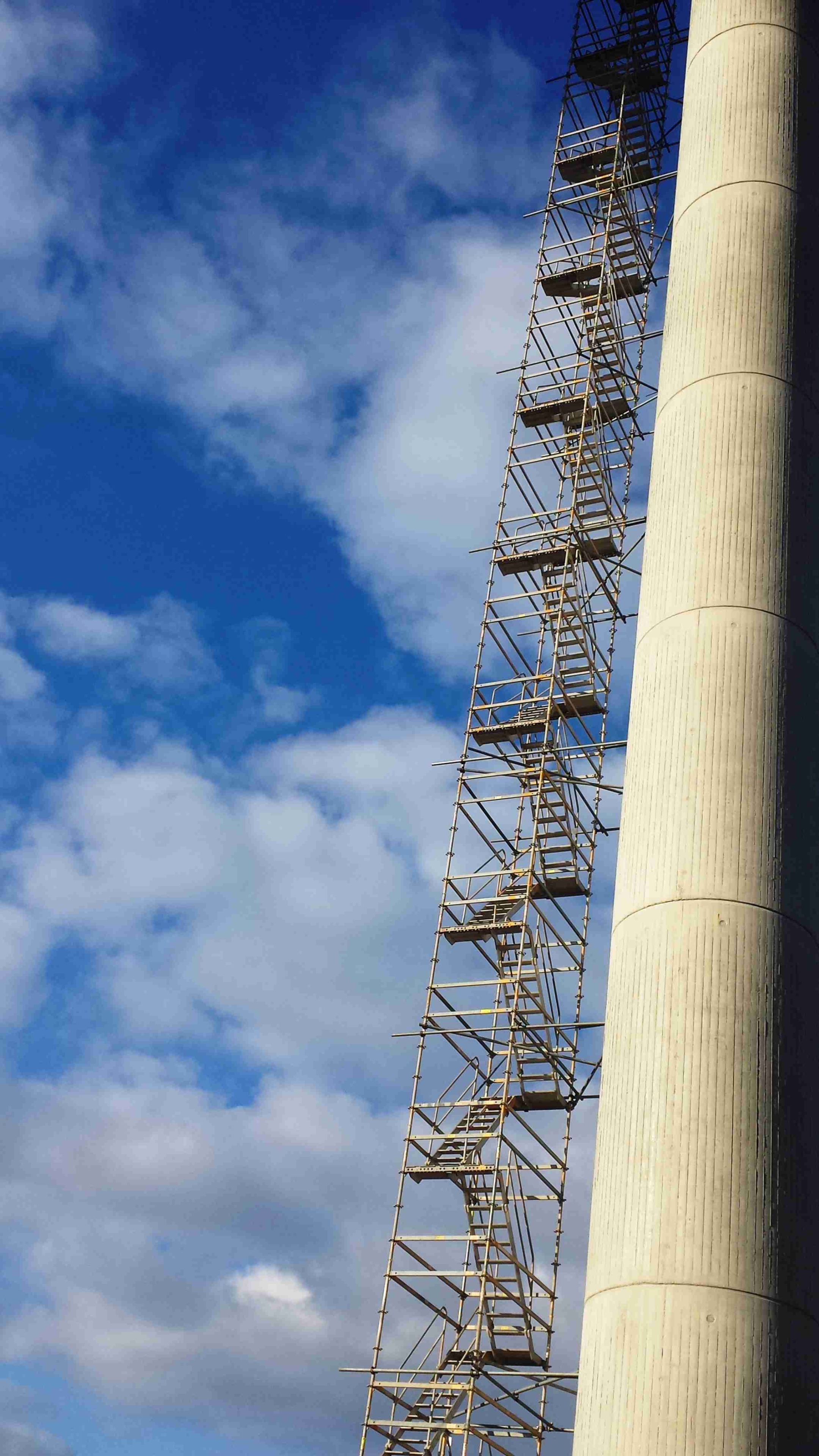 Vor einem frühlingshaften Wolkenhimmel führt eine Leiter außen an einem hohen Kamin in die Höhe.