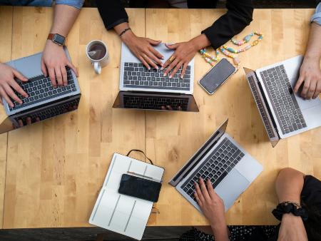 Teamwork (c) Louise Viallesoubranne/unsplash Blick auf Tischplatte, Menschen haben darauf ihre Laptops aufgeklappt und die Hände auf der jeweiligen Tastatur. Da die Hände unterschiedliche Hautfarben haben, dient das Bild zur Illustration des Themas 'Rassismus begegnen' im beruflichen Alltag.
