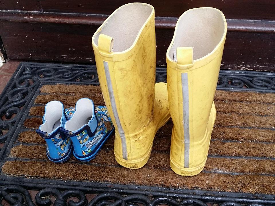 Nebeneinander auf einer Fußmatte steht ein Paar Gummistiefel eines Kindes (blau) neben einem Paar für Erwachsene (gelb).