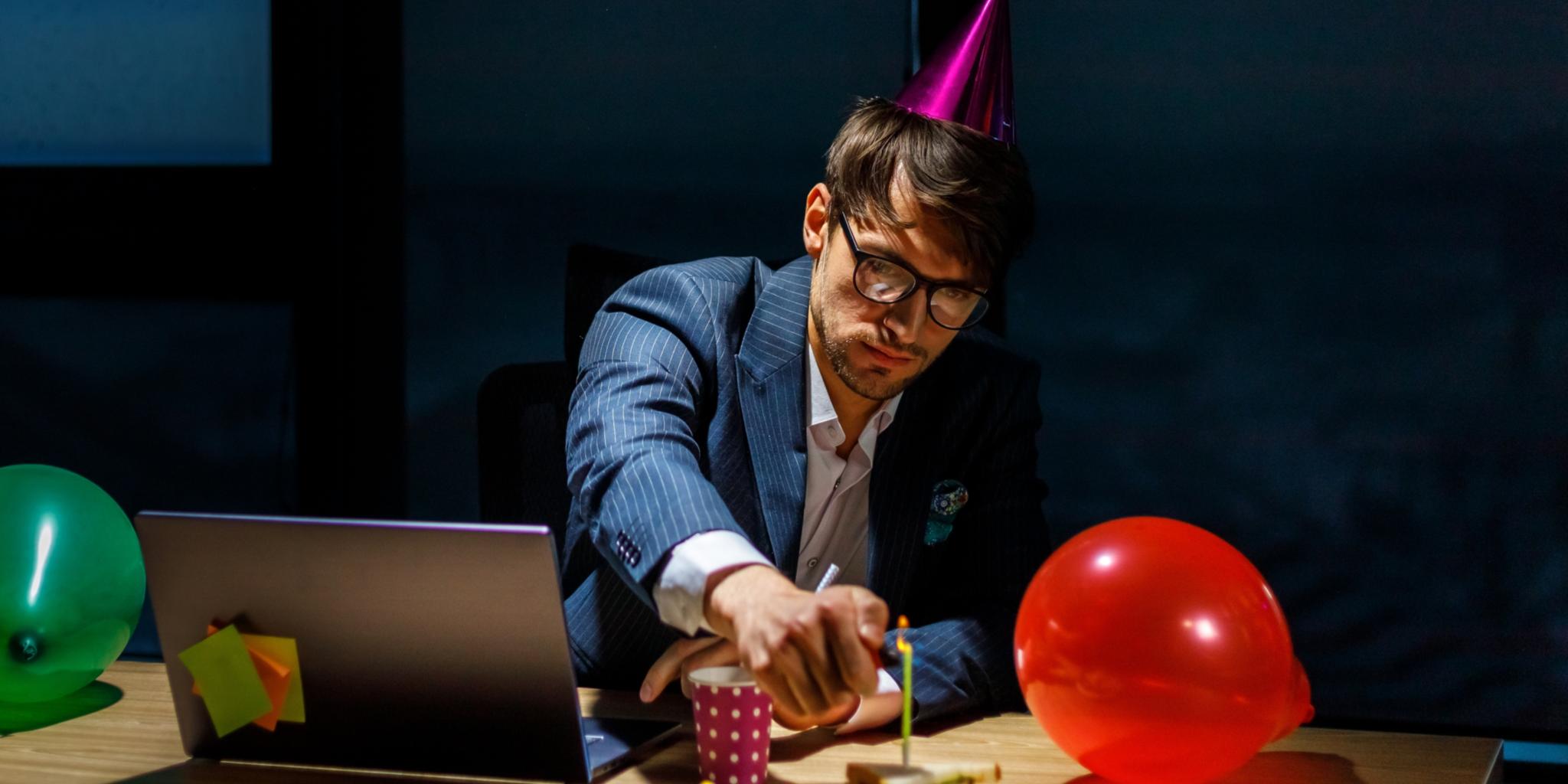 Ein Chef sitzt mit Partyhut alleine vor dem Laptop, neben ihm Luftballons, er zündet eine Kerze auf einem Geburtstagskuchen an.