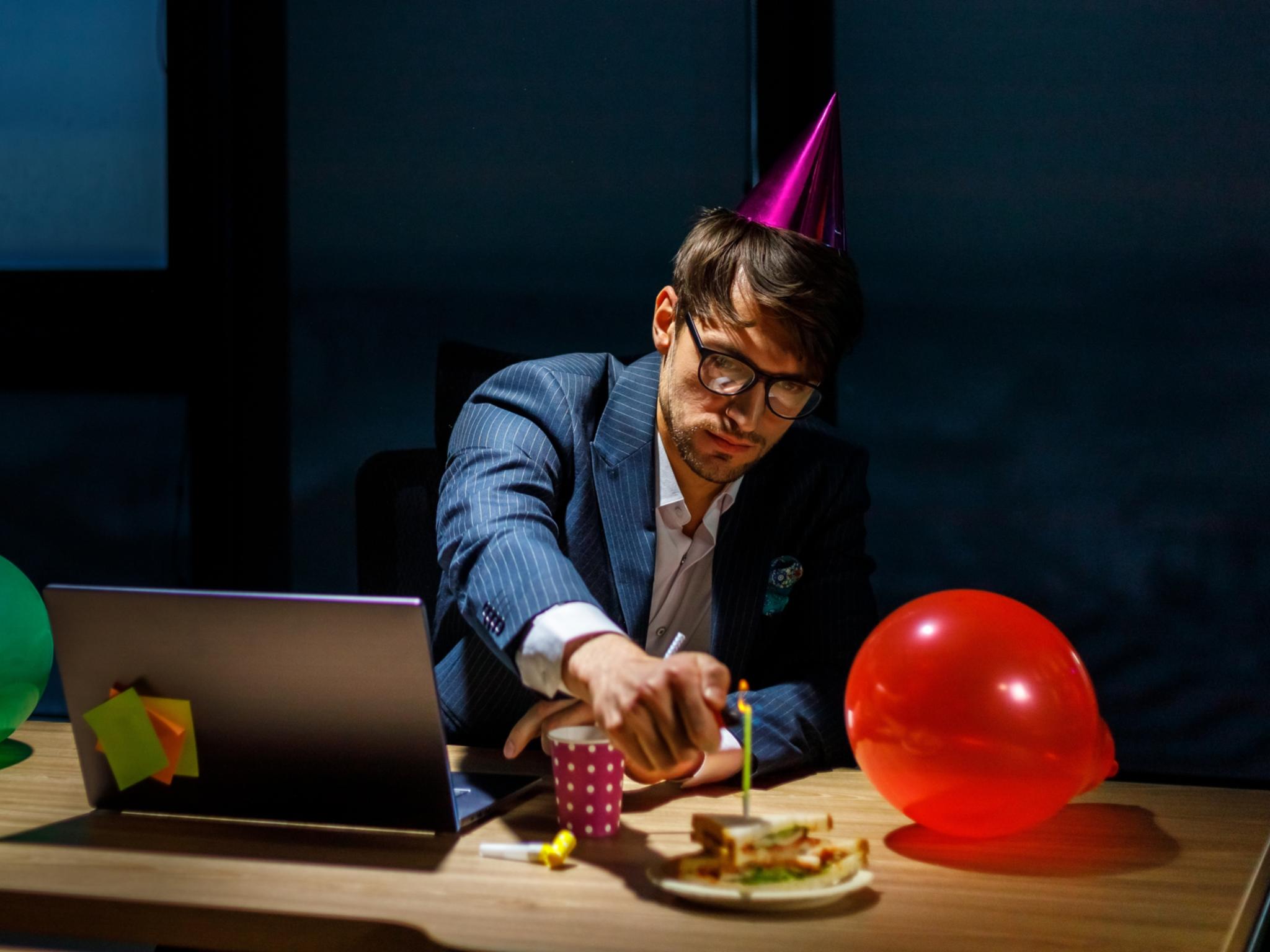 Ein Chef sitzt mit Partyhut alleine vor dem Laptop, neben ihm Luftballons, er zündet eine Kerze auf einem Geburtstagskuchen an.