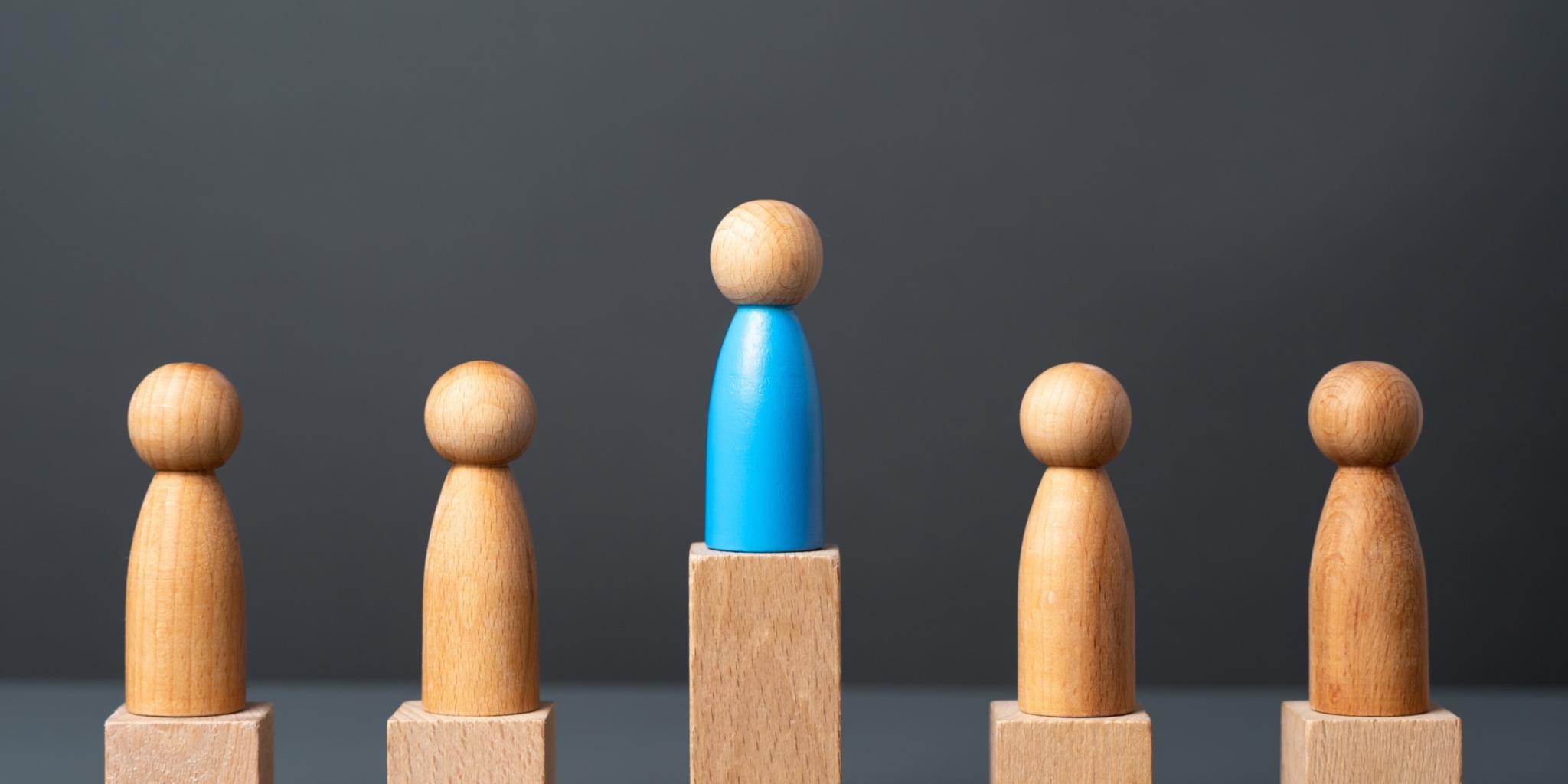 Fünf Holzspielfiguren auf kleinen Holzpodesten, die mittlere Figur steht auf einem höheren Podest, sie überragt die anderen.