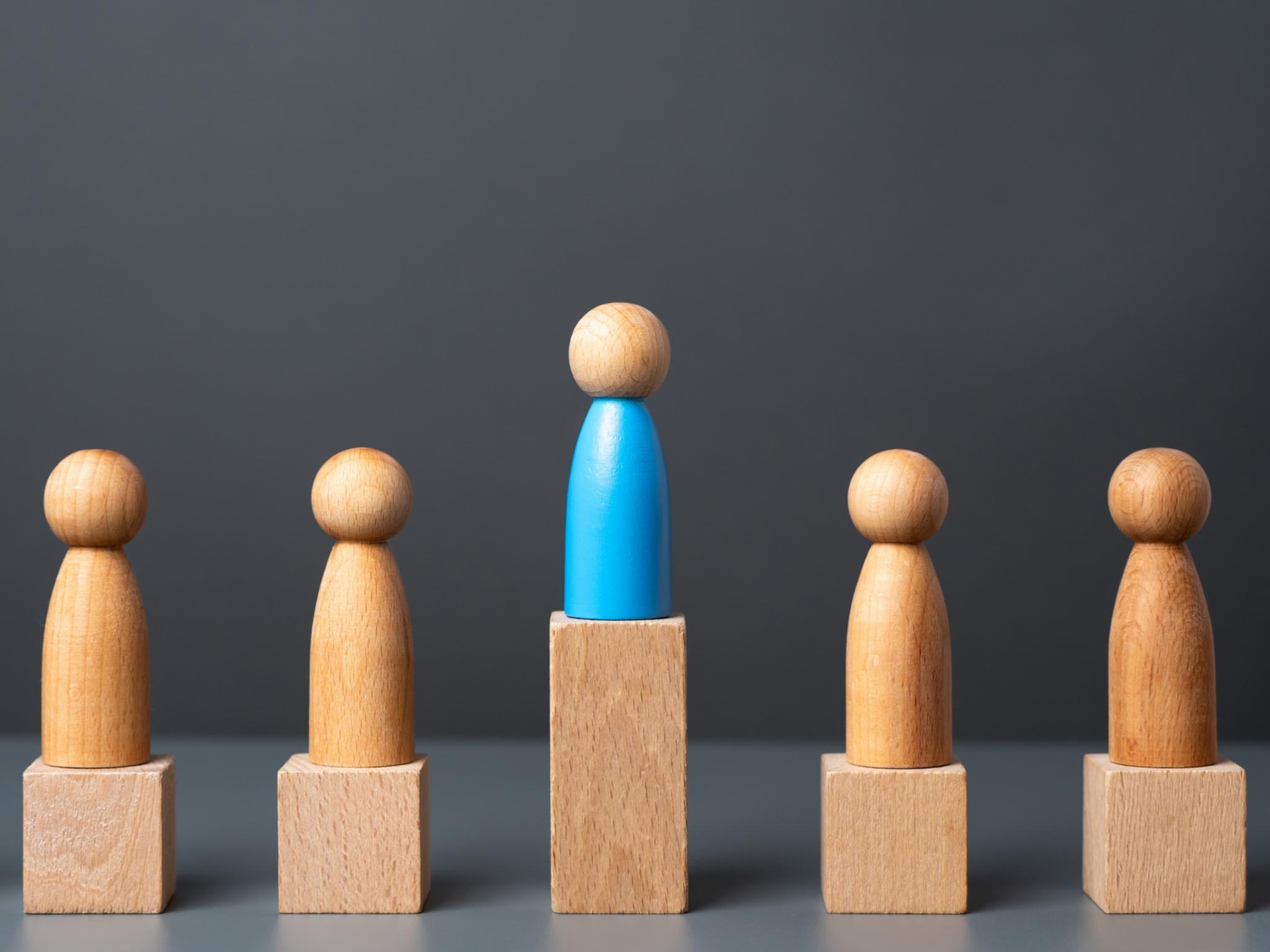Fünf Holzspielfiguren auf kleinen Holzpodesten, die mittlere Figur steht auf einem höheren Podest, sie überragt die anderen.