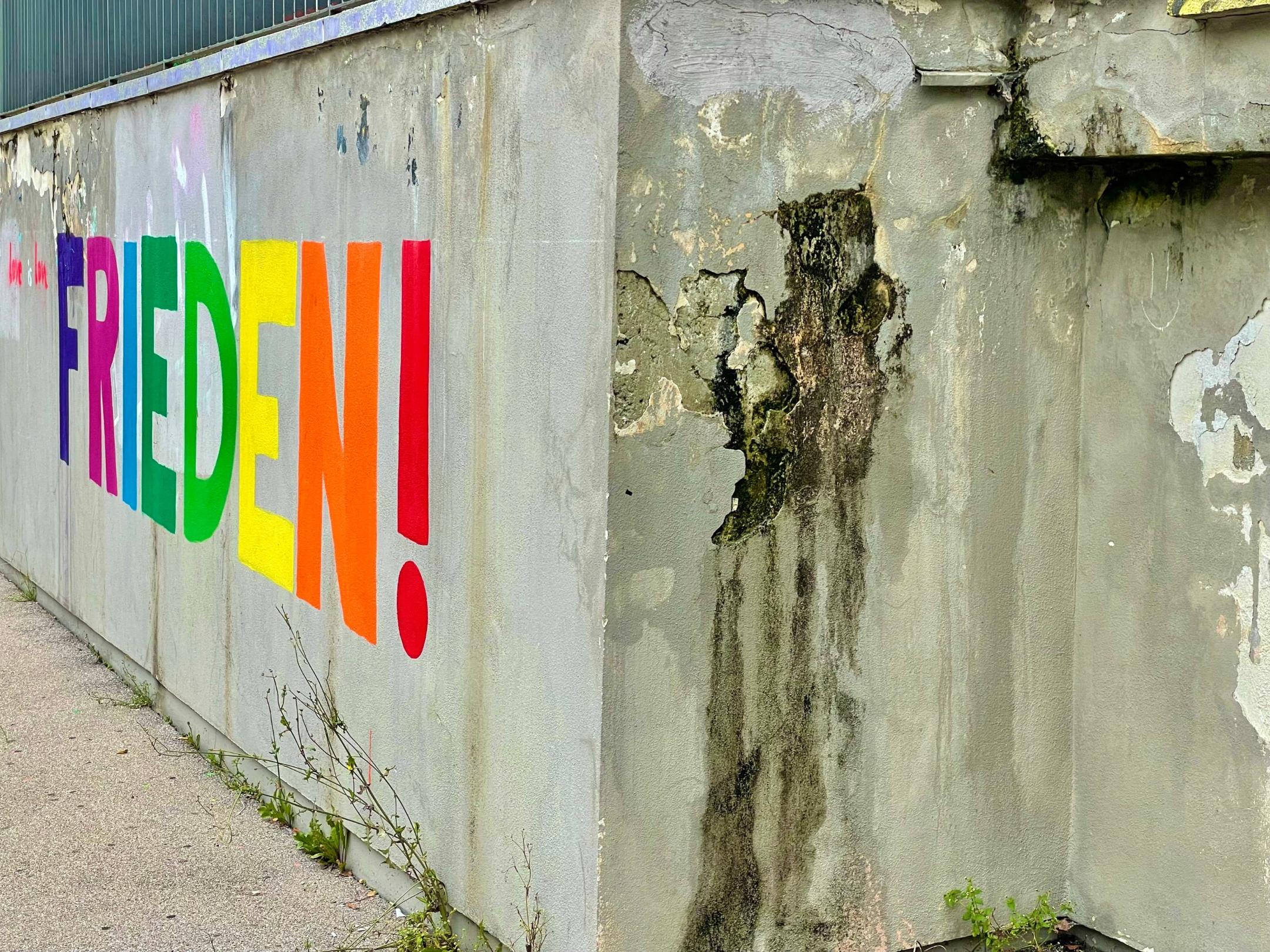 Eine Betonwand, daran ist Großbuchstaben das Wort 'FRIEDEN' mit Ausrufezeichen angemalt.