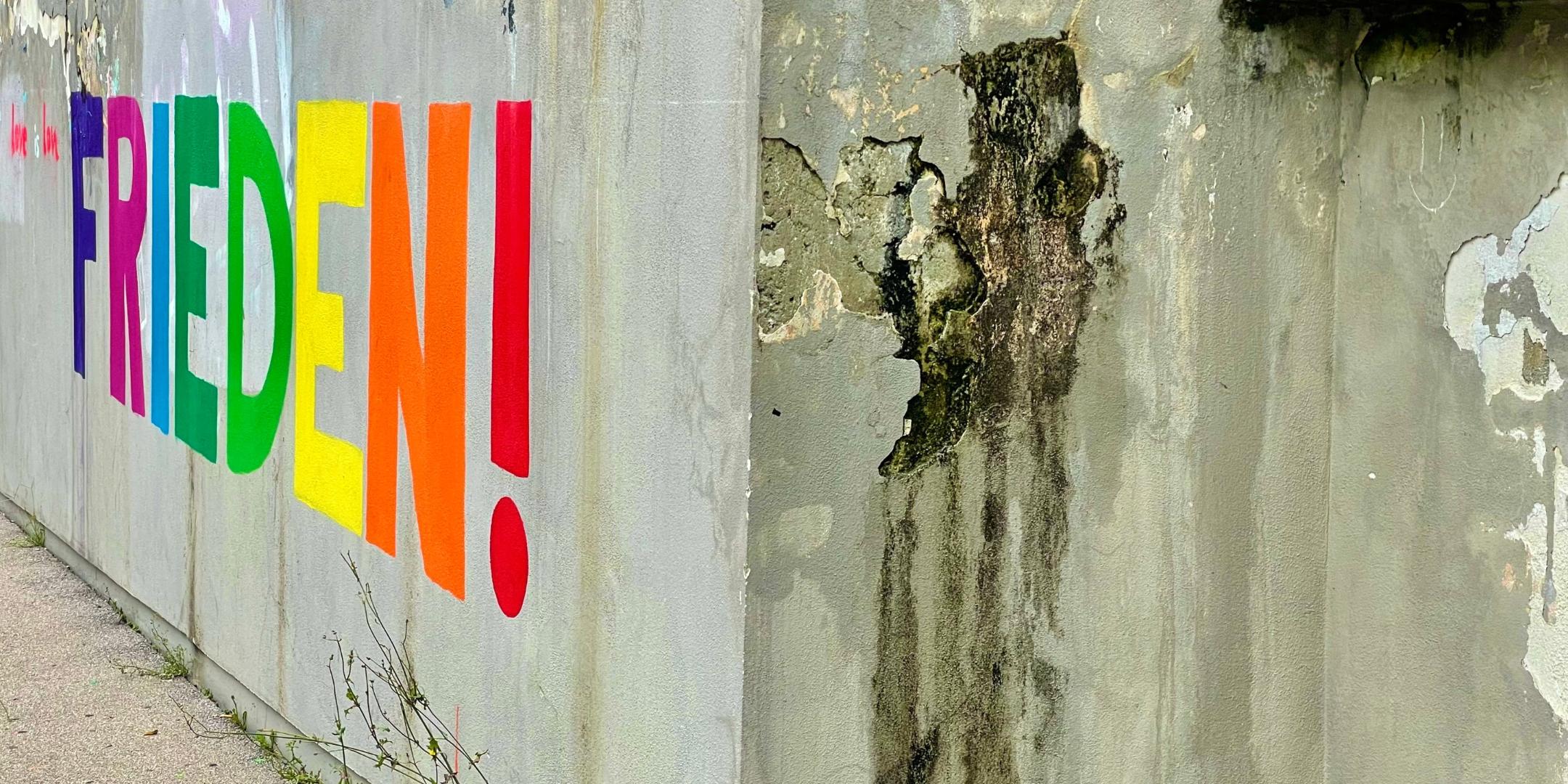 Eine Betonwand, daran ist Großbuchstaben das Wort 'FRIEDEN' mit Ausrufezeichen angemalt.
