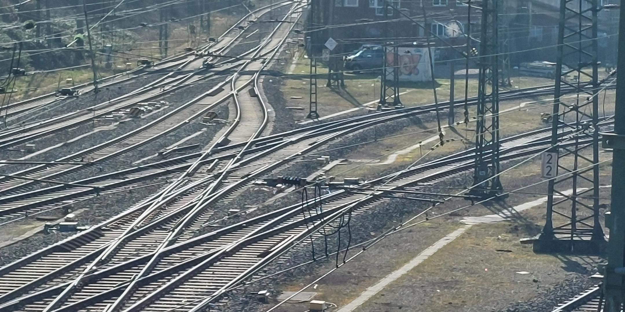 Blick von einer Eisenbahnbrücke auf Bahngleise, im Hintergrund Häuser und bewaldete Hügel.
