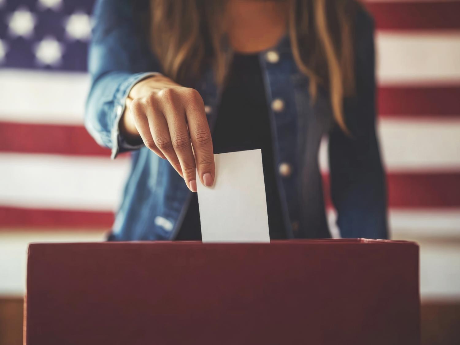 Eine Frau steckt einen Wahlzettel in eine Wahlurne, das Gesicht ist nicht erkennbar. Hinter ihr die Flagge der USA.